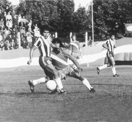 Maradona de adolescente y larga cabellera disputando un balón en el juego entre Argentinos, camiseta con franja cruzada, y Talleres, camiseta a rayas verticales, en 1976 Maradona de adolescente y larga cabellera disputando un balón en el juego entre Argentinos, camiseta con franja cruzada, y Talleres, camiseta a rayas verticales, en 1976